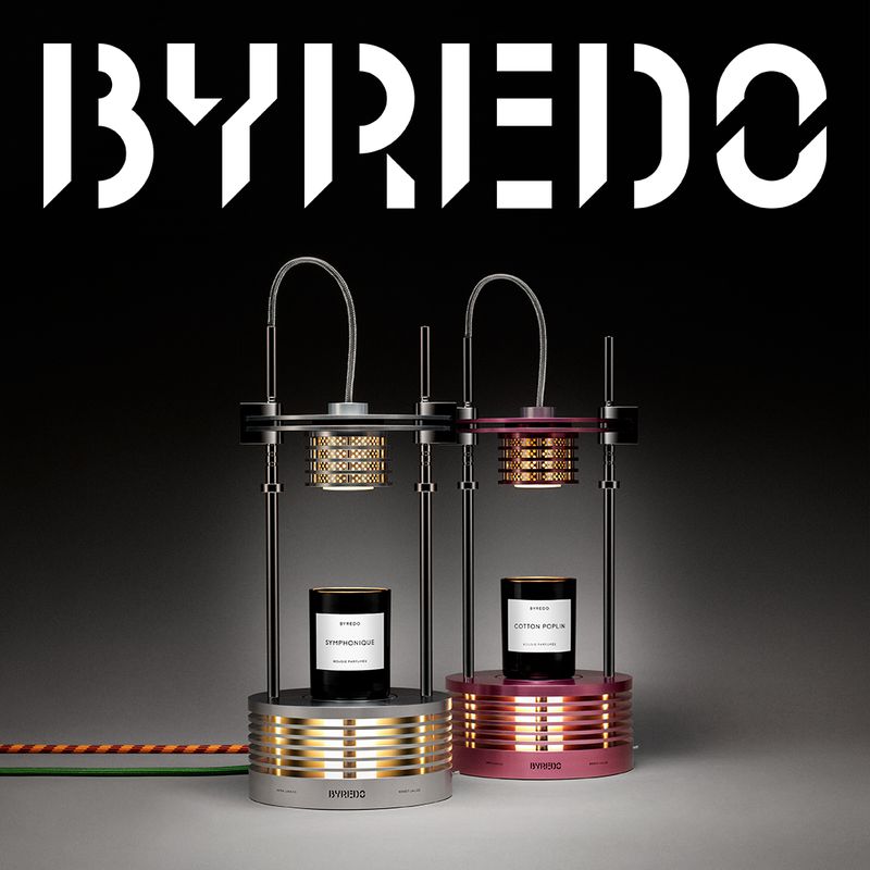 Two lit Byredo lamps with perfume stand on a gray surface against a dark background, showcasing luxury home fragrance products.