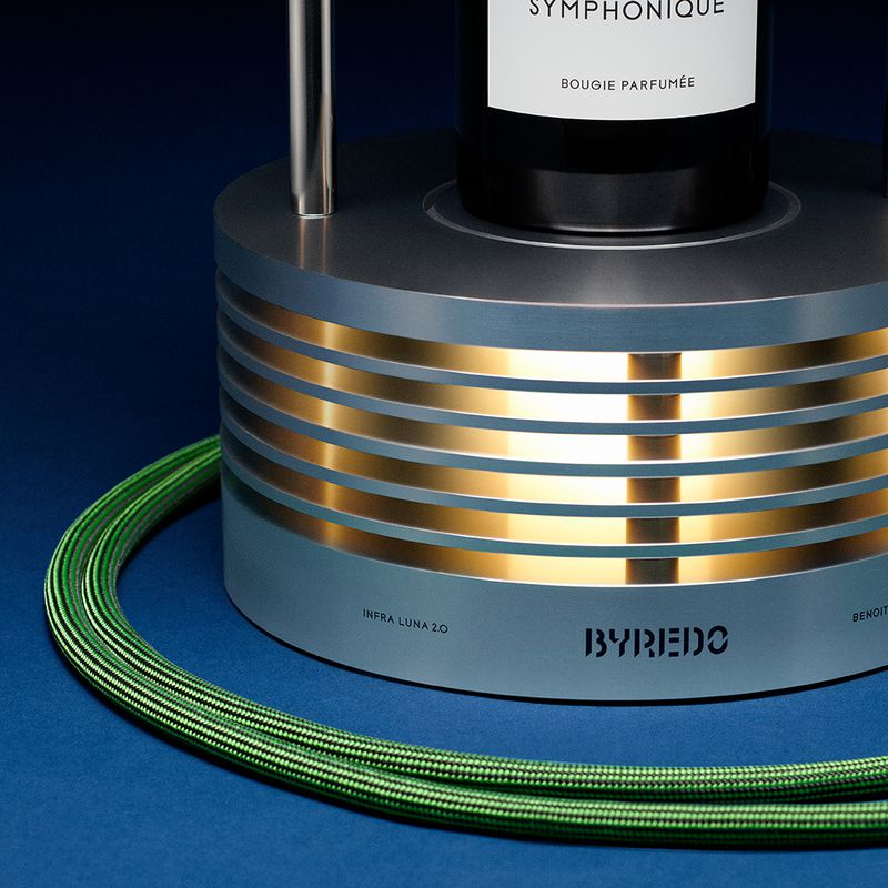 Close-up studio shot of a Byredo Symphonique candle on a brushed metal warmer with gold accents and a coiled green power cord.