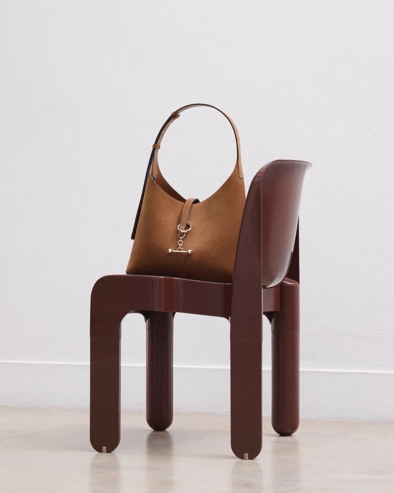 Brown suede hobo bag with gold hardware on brown modernist chair against a white wall and floor in a brightly lit photo studio.