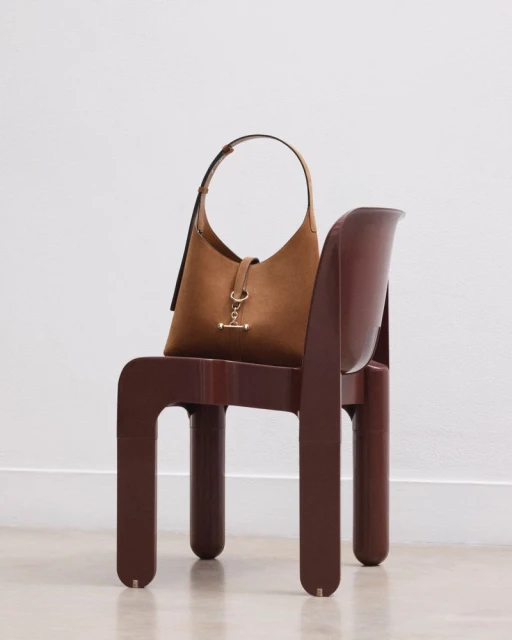 Brown suede hobo bag with gold hardware on brown modernist chair against a white wall and floor in a brightly lit photo studio.