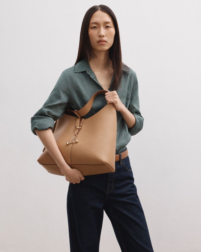 Adult woman poses indoors holding tan leather handbag, wearing a teal button-down shirt, with long dark hair and a light complexion.