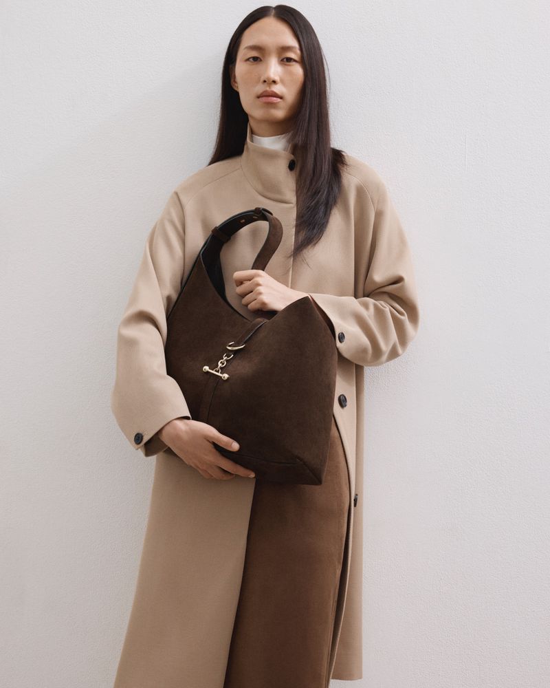 An Asian woman in a tan coat holds a large brown suede hobo bag in a studio setting, showcasing fashion and design.