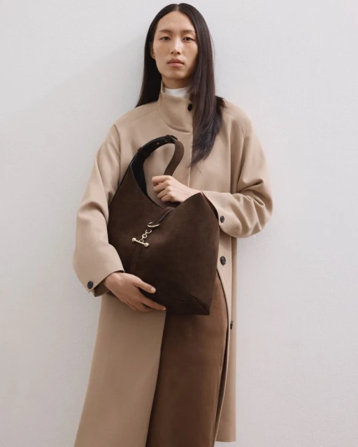 An Asian woman in a tan coat holds a large brown suede hobo bag in a studio setting, showcasing fashion and design.