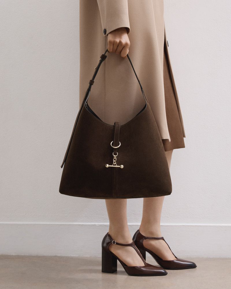 A person wearing a beige coat and brown T-strap heels holds a large brown suede hobo bag with metal hardware in a studio setting.