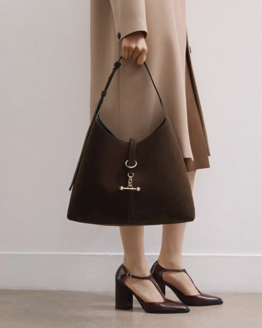 A person wearing a beige coat and brown T-strap heels holds a large brown suede hobo bag with metal hardware in a studio setting.