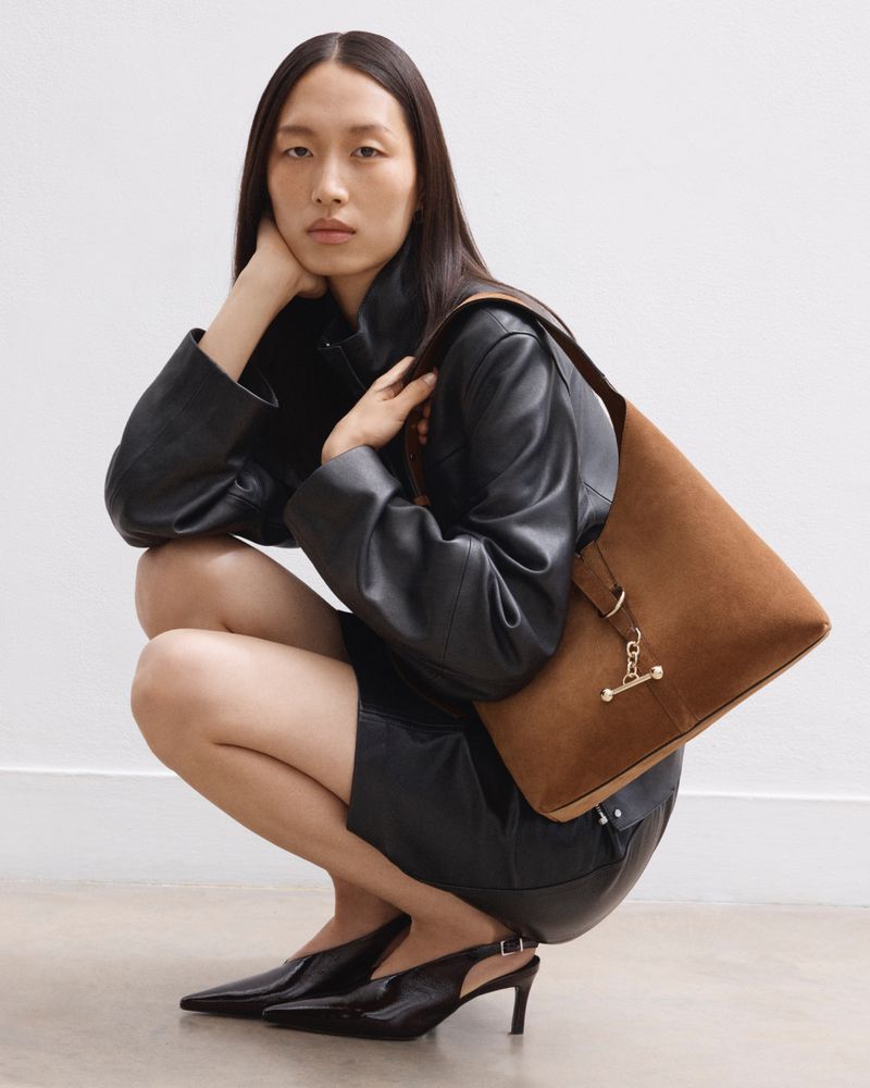 An Asian woman squats indoors wearing a black leather jacket, skirt and heels. A brown suede purse hangs from her shoulder.