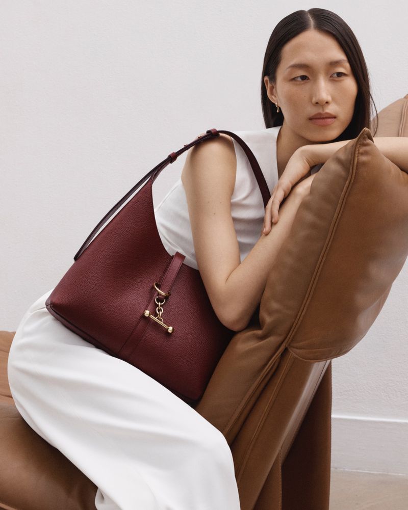 Elegant woman poses with a maroon leather purse in a minimalist indoor fashion shot, highlighting style and quality.