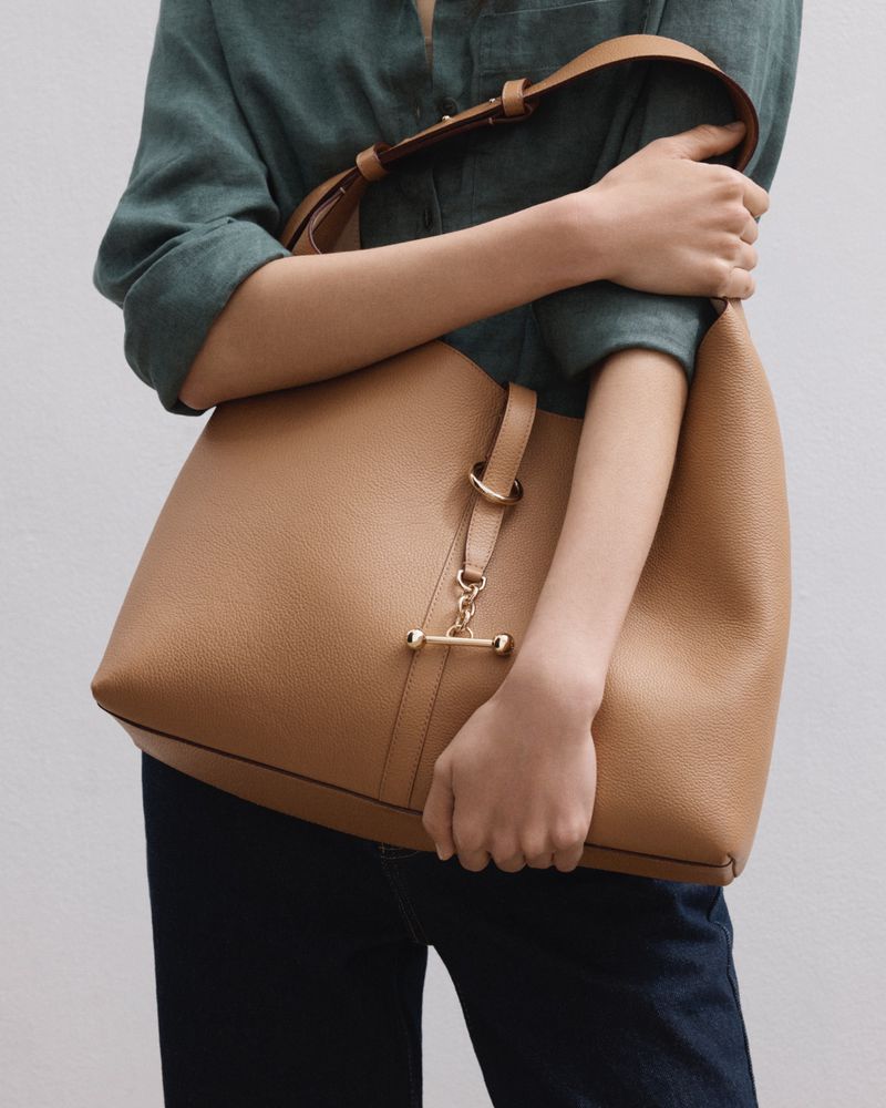 Person holding a large tan leather tote bag in a studio shot. The bag features a top handle, shoulder strap, and gold metal hardware.