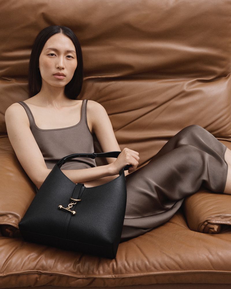 Woman poses with a black leather handbag while reclining on a brown leather sofa. The bag features gold hardware.