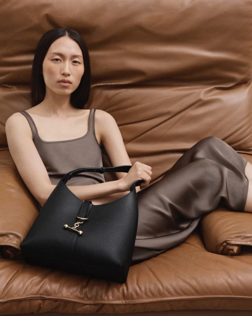Woman poses with a black leather handbag while reclining on a brown leather sofa. The bag features gold hardware.