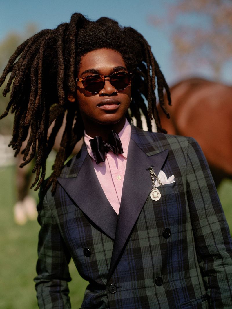 A stylish individual with dreadlocks is posing outdoors. They are wearing sunglasses, a pink shirt, and a patterned blazer with a bow tie.