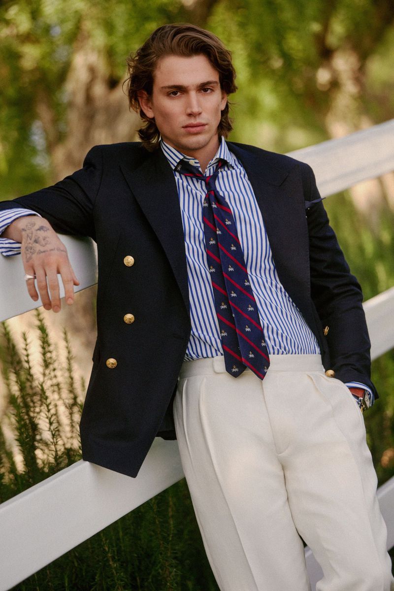 Adult male in a blue and white striped shirt, blue blazer with gold buttons, and white pants stands outdoors, leaning on a white fence.