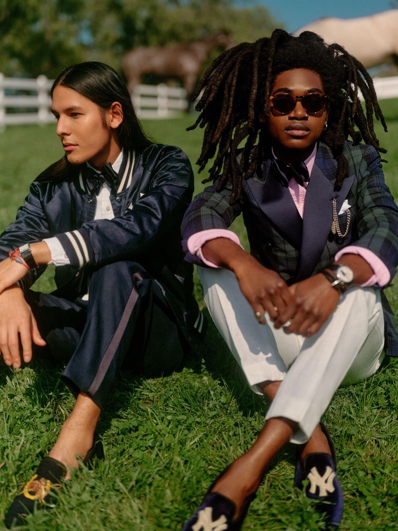 Two stylish men sit in a grassy field with a horse and white fence in the background, showcasing fashion and outdoor lifestyle.