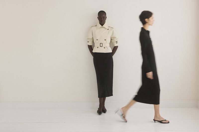 Two women pose in a fashion shot in front of a white wall, one in a jacket and skirt, the other in a dress and motion blur.