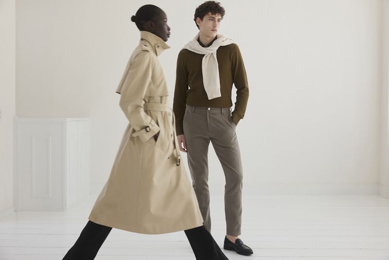 A stylish pair, one in a beige trench coat and the other in brown tones, pose against a bright backdrop, showcasing fall fashion.