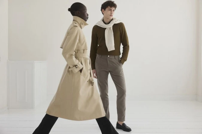 A stylish pair, one in a beige trench coat and the other in brown tones, pose against a bright backdrop, showcasing fall fashion.