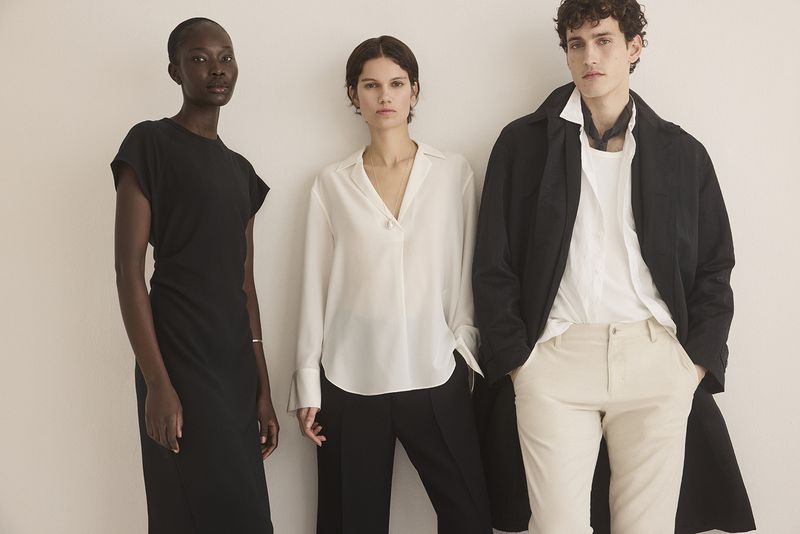 Three models pose in a studio against a neutral backdrop. One wears a black dress, another a white blouse and black pants, and the third a coat.