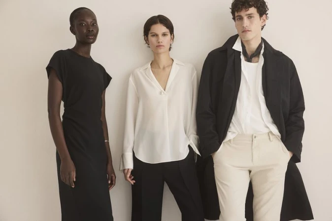 Three models pose in a studio against a neutral backdrop. One wears a black dress, another a white blouse and black pants, and the third a coat.