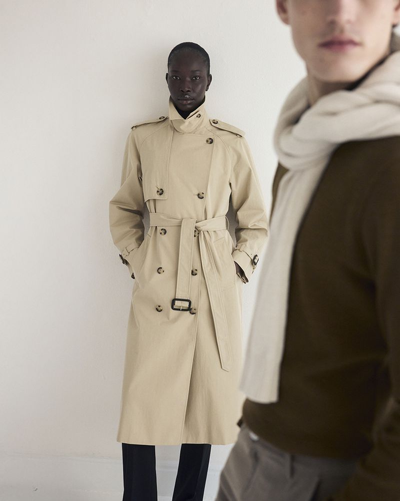 Two fashion models in studio wearing coats and scarves. One Black woman stands in front of a light background and a white man stands blurred.