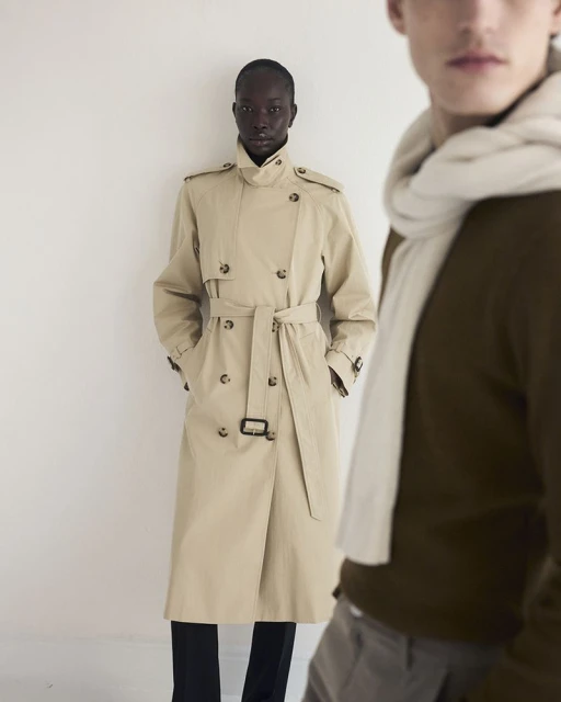 Two fashion models in studio wearing coats and scarves. One Black woman stands in front of a light background and a white man stands blurred.