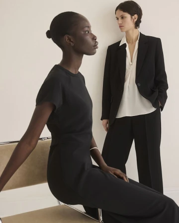 Two women in a neutral indoor setting. One woman with dark skin sits on a chair in a black dress while another woman stands in a blazer.