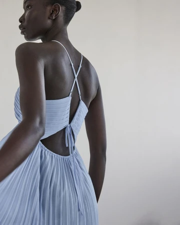 Back view of an African American woman in a light blue pleated dress with thin straps, indoors against a light background.