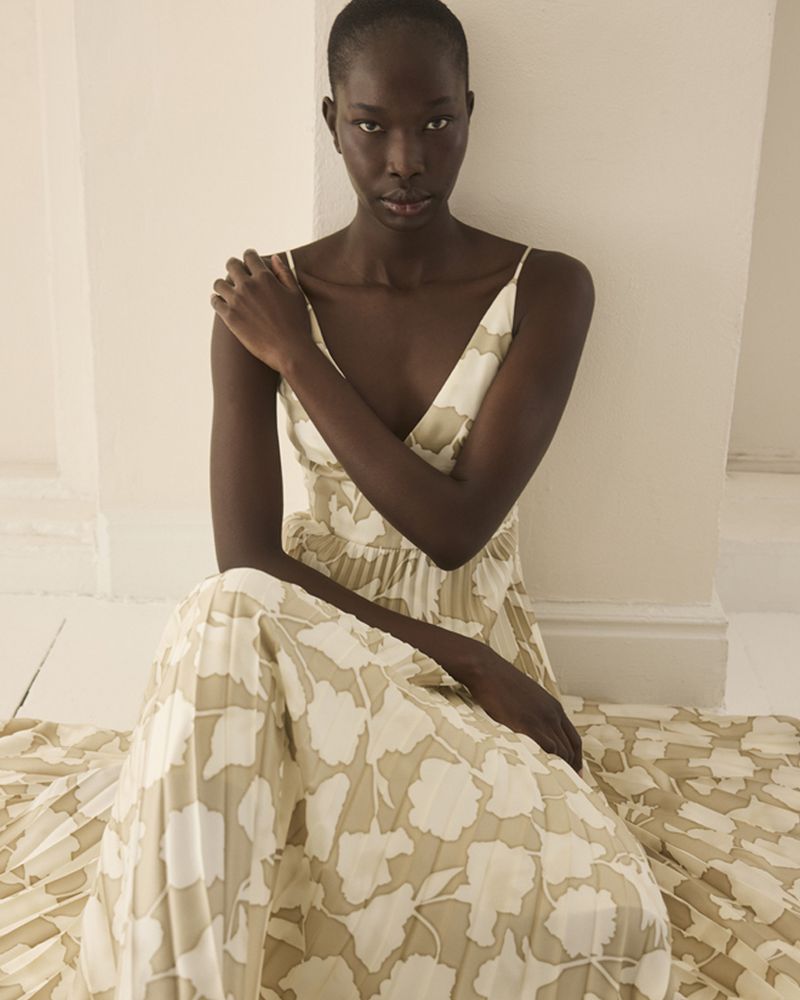 Dark-skinned woman in floral print dress poses indoors. Model is sitting and looking at the camera with her arm on her shoulder.