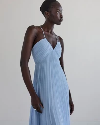Model Adhel Bol wearing a light blue pleated maxi dress against a neutral white background for Club Monaco spring 2025.