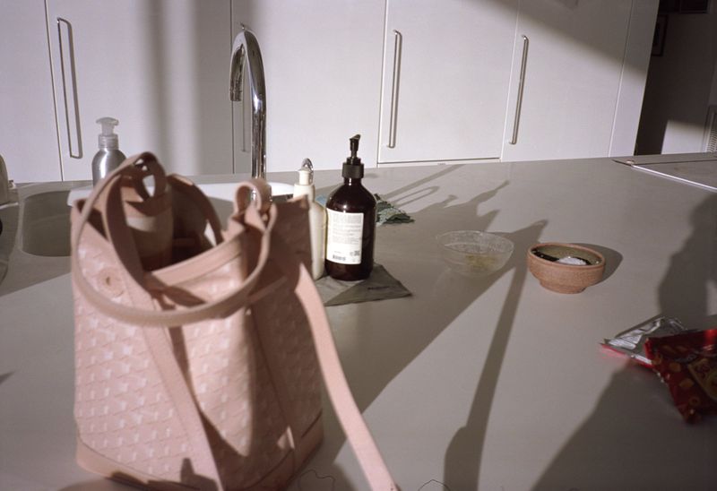 Light pink tote bag on a white countertop with a sink, bottles, and bowls, casting long shadows in a bright indoor kitchen setting.