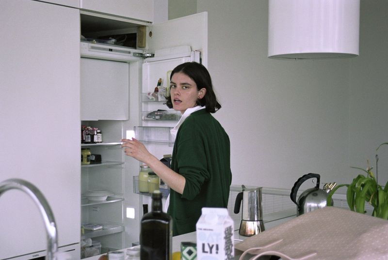 Young woman in a green sweater looks into an open refrigerator in an indoor kitchen setting. An array of kitchen objects are on the counter.