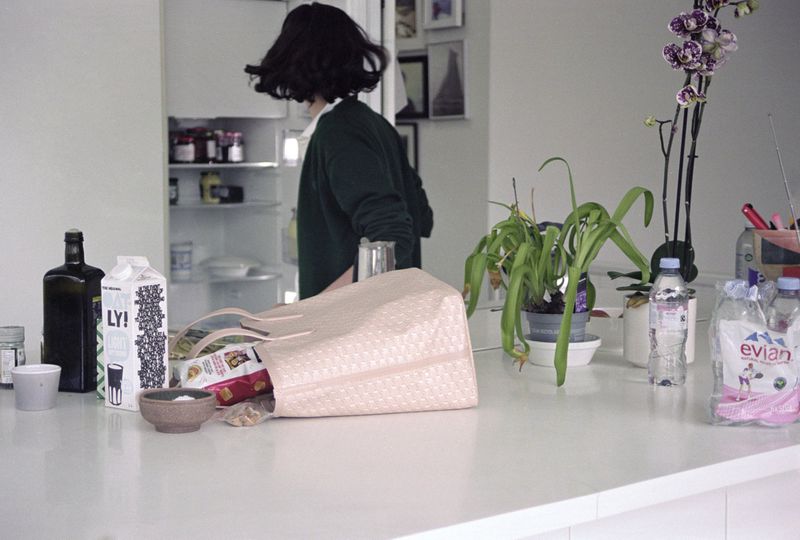 A woman stands by an open refrigerator in a bright kitchen filled with an assortment of groceries and household objects on the counter.