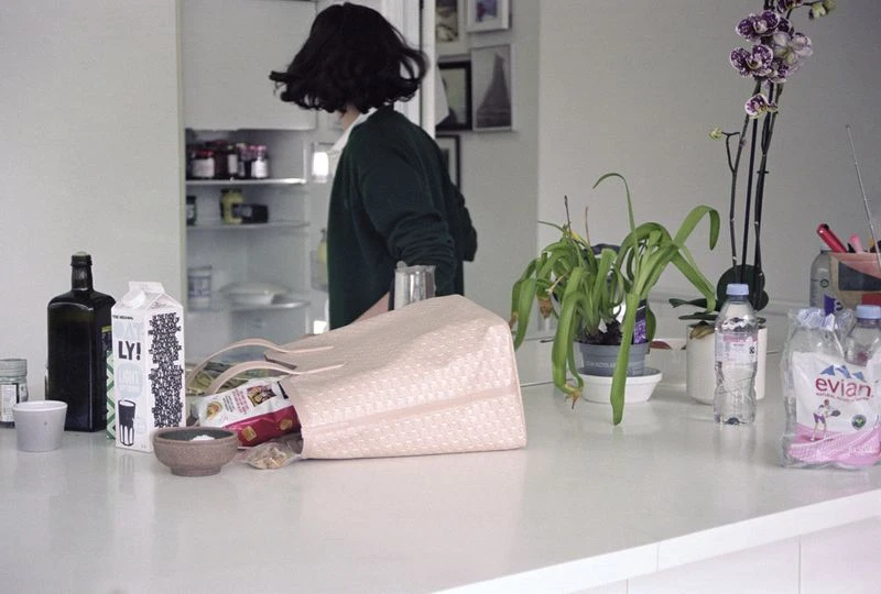 A woman stands by an open refrigerator in a bright kitchen filled with an assortment of groceries and household objects on the counter.