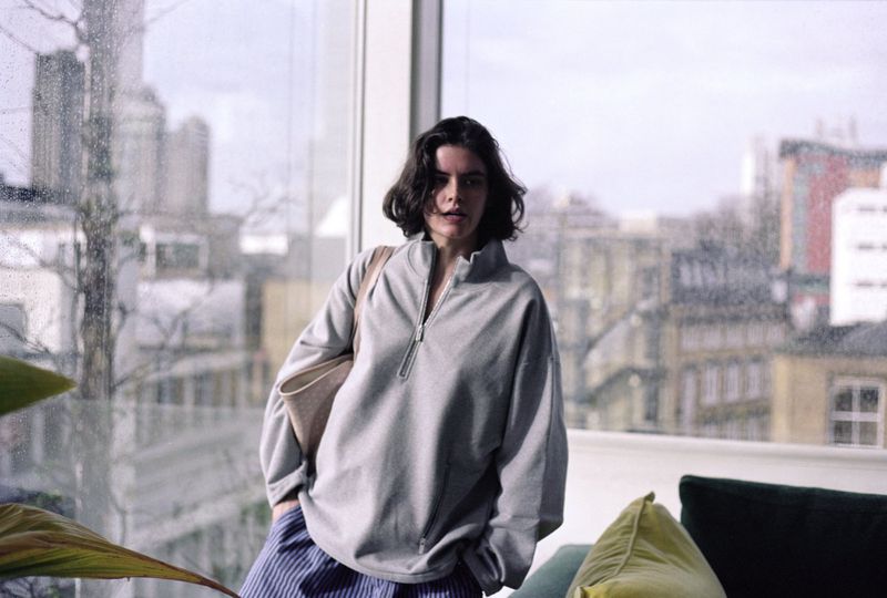 Woman in gray sweatshirt and blue striped pants stands indoors by a window with city view; she carries a tan tote bag over her shoulder.
