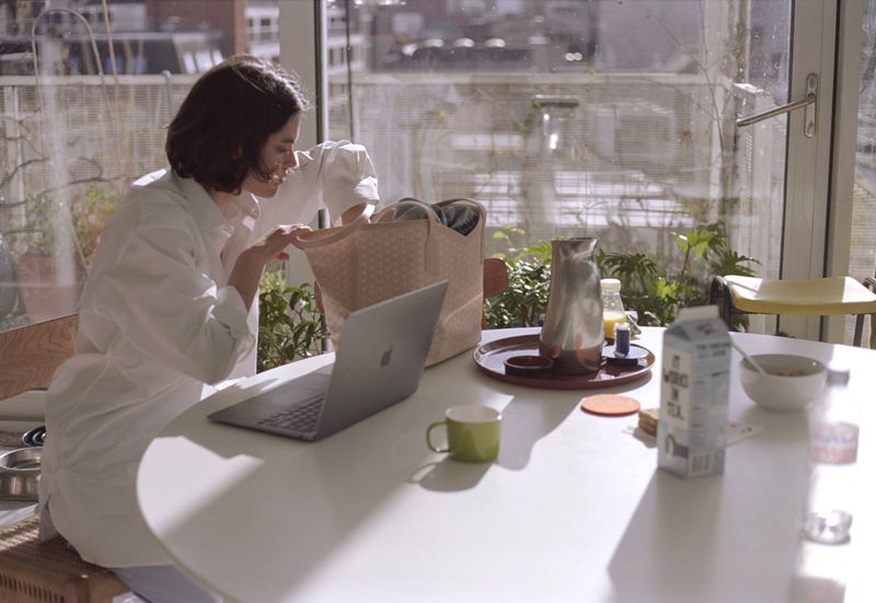 Adult woman sits at white table indoors with laptop and tote bag near sunny window with city view, enjoying breakfast with a carton of milk.