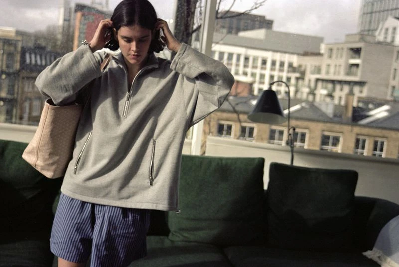 Adult female in a grey pullover sweater and blue striped shorts indoors near a window with city views. Carrying a beige tote bag.