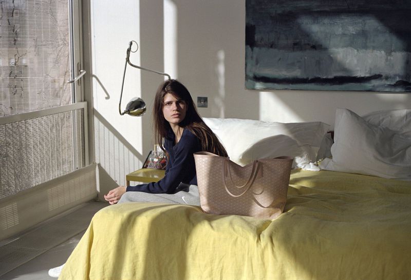 A woman sits on a yellow bed with a patterned beige bag, indoors with natural light and a painting on the white wall behind.