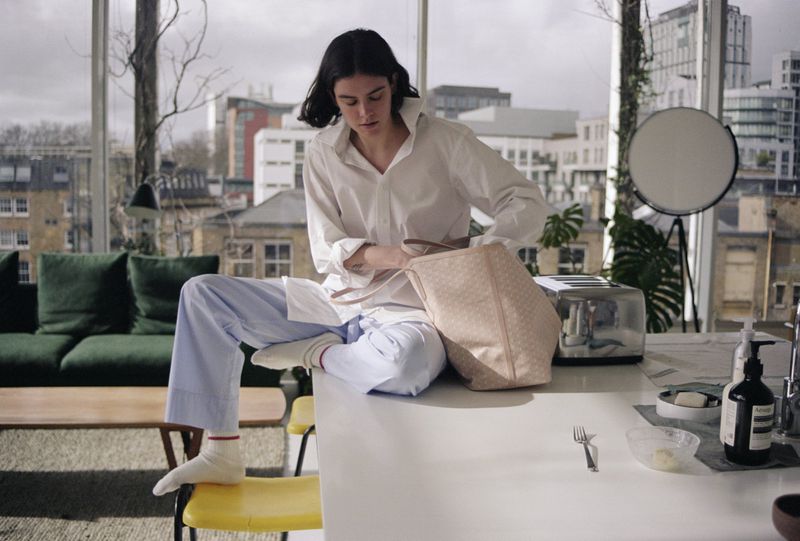 Woman in oversized shirt and pajama pants sits on a table with a purse in an apartment with a city view from a large window.