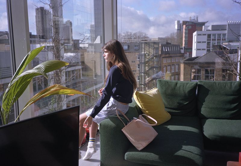 A young woman in a navy sweatshirt sits on a green couch near a window overlooking an urban city, beside a houseplant and pink tote bag.