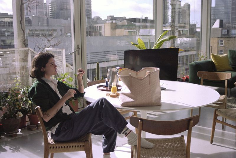 Indoor apartment shot of a woman seated near a table with food, drinks, and a tote bag, with a cityscape visible through a window.