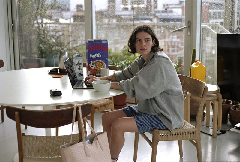 Young adult woman with short brown hair sits indoors at a table using a laptop with a cereal box, pitcher, and bowl nearby.