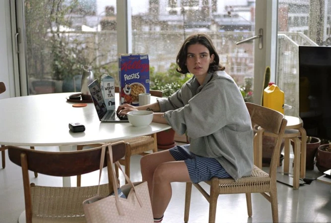 Young adult woman with short brown hair sits indoors at a table using a laptop with a cereal box, pitcher, and bowl nearby.