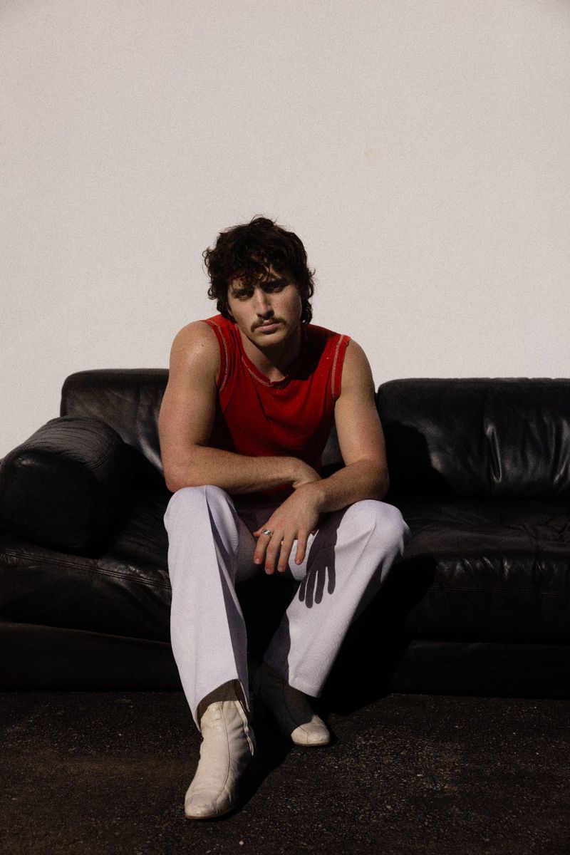 Adult male with mustache sits on black leather couch in a red tank top and white pants in front of a white wall in a studio.