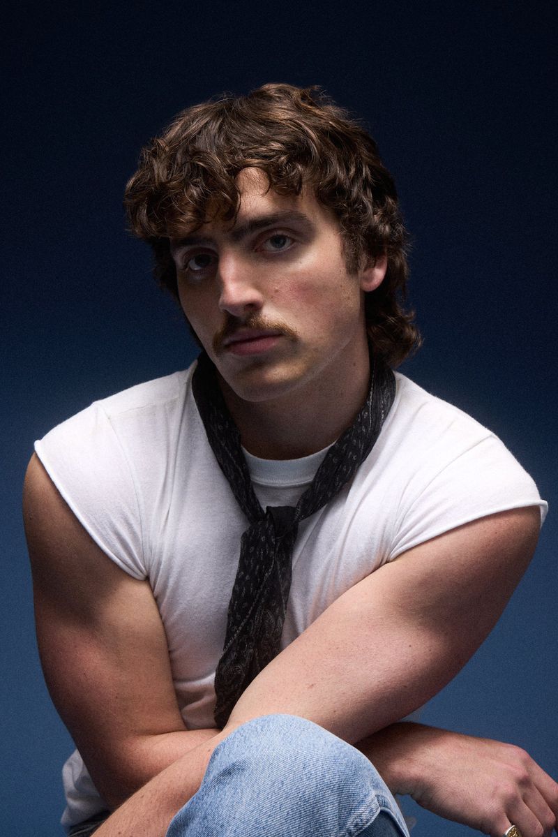 Portrait of a young adult male with curly brown hair, wearing a white t-shirt and a patterned tie, posing against a dark blue backdrop.