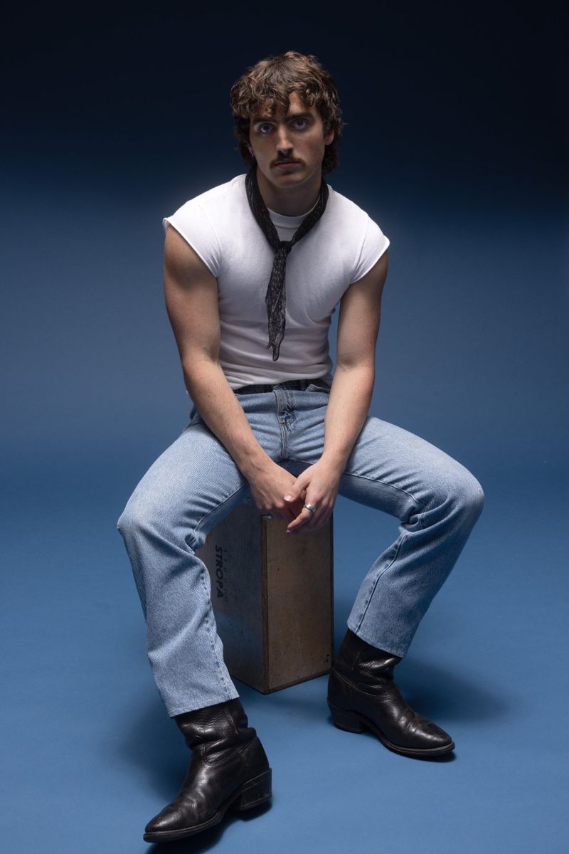 Portrait of a young adult male with dark hair sitting on a box, wearing a white t-shirt, light blue jeans, and black boots against a blue background.