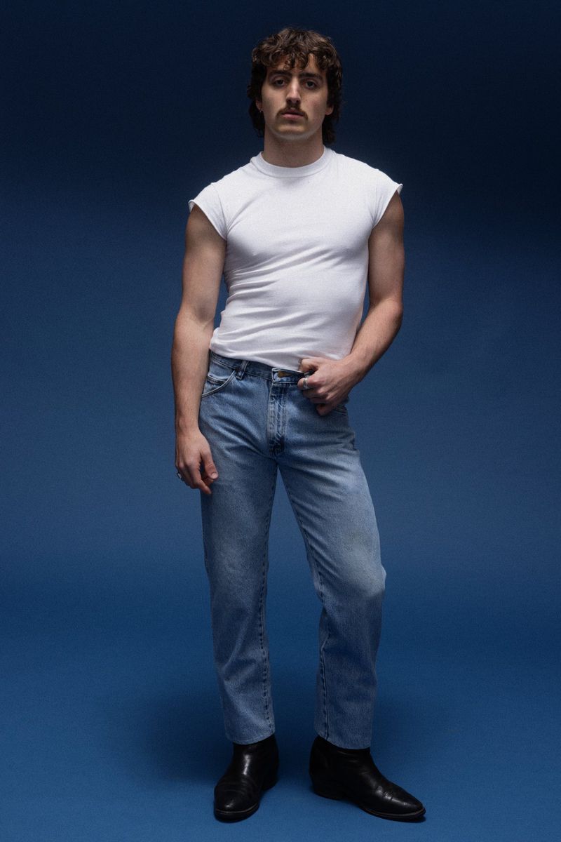 Full-length portrait of a man in a white sleeveless t-shirt and jeans standing against a blue background in a photo studio setting.