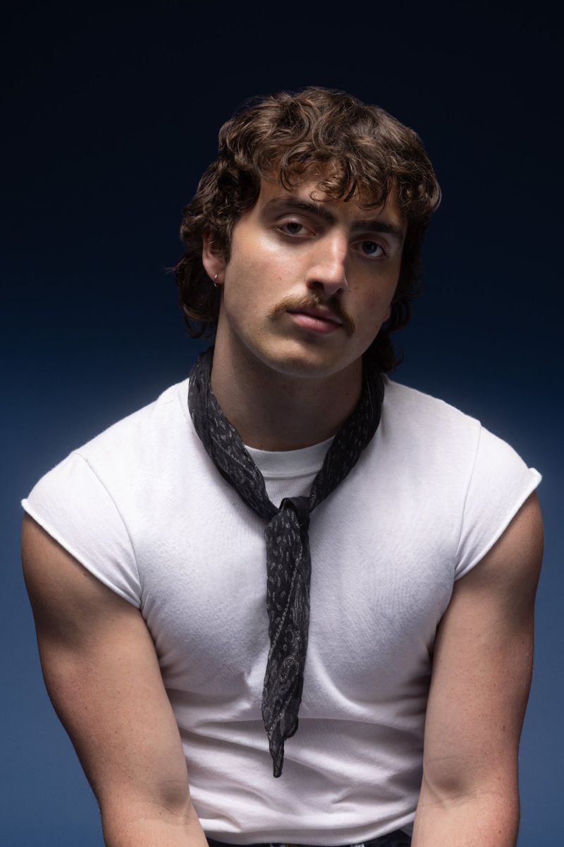 Portrait of a young man with a mustache wearing a white t-shirt and a patterned scarf against a dark blue background, posing in the photo studio.