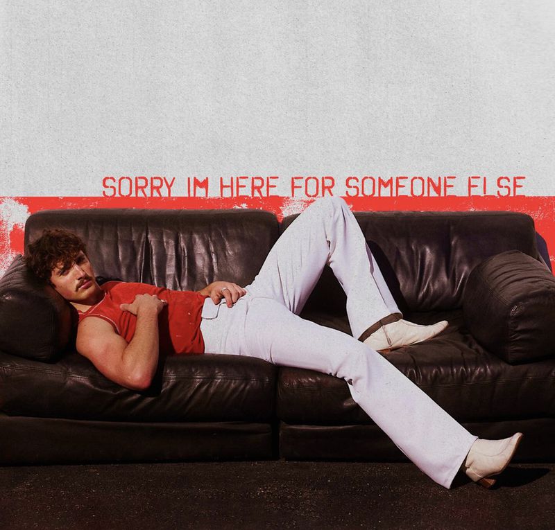 An adult male with a mustache, wearing a red shirt and white pants, lounges on a black leather couch against a painted wall.