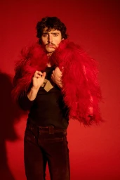 Musician Benson Boone posing in a vibrant red feathered coat against a matching red studio background.