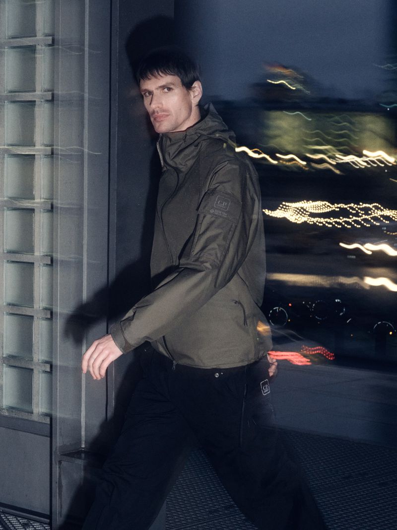 Man in olive green hooded jacket and black pants walks near a grey wall at night with blurred lights in the background.