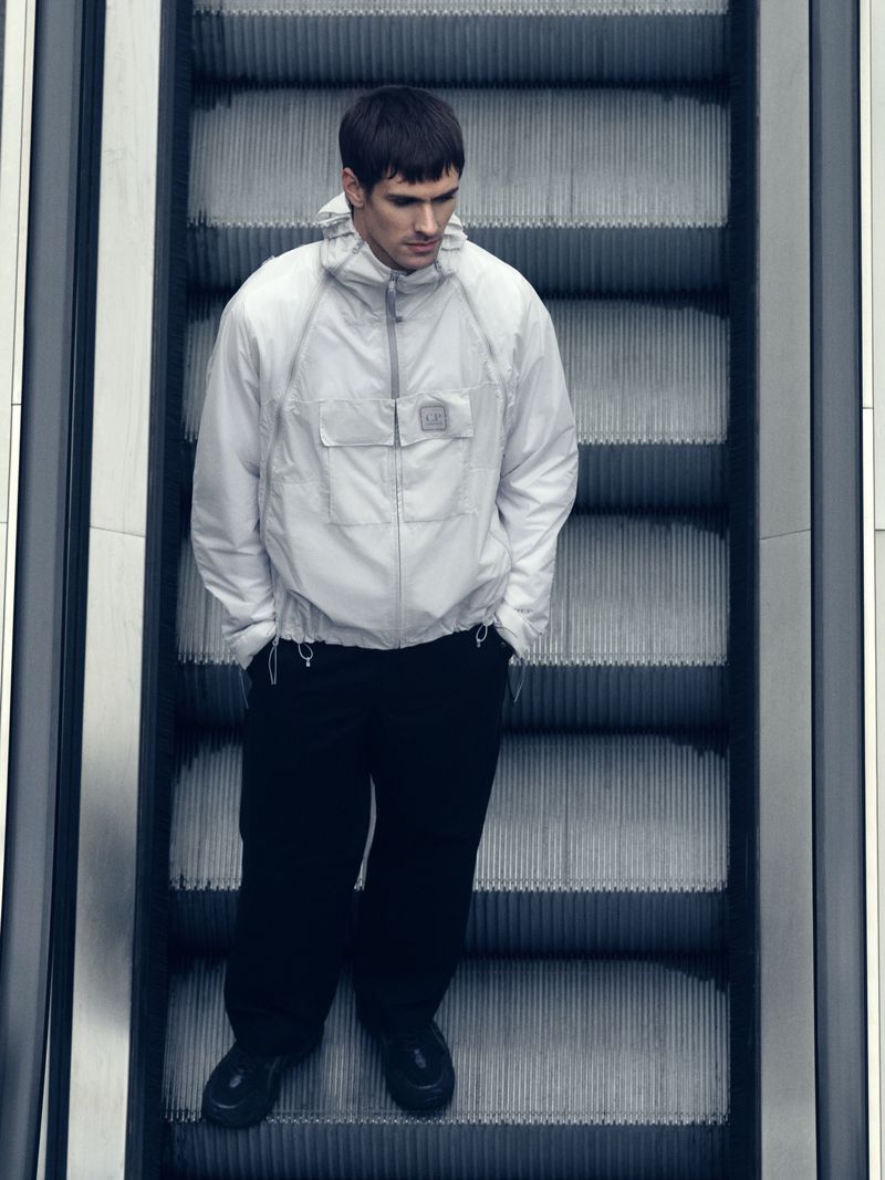 Man in a white jacket standing on a gray escalator. The jacket has a hood. He is wearing black pants and shoes.